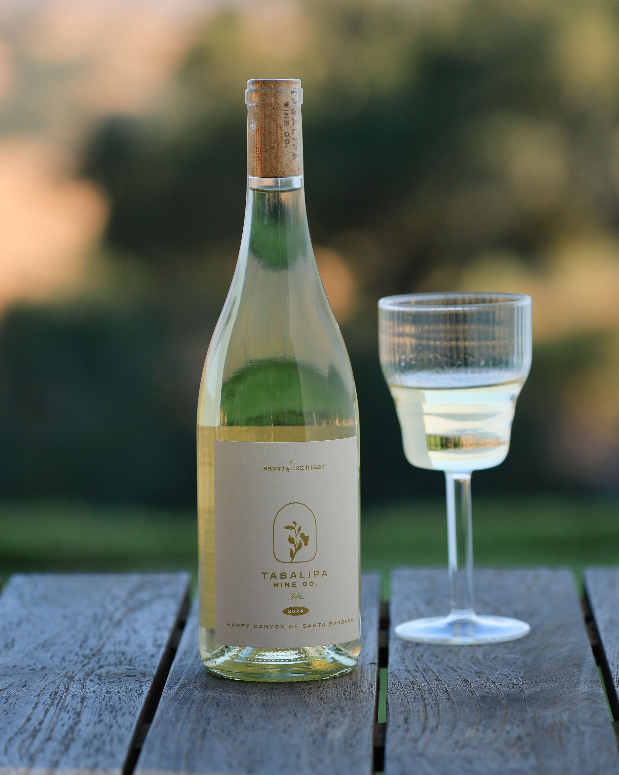 A bottle of Tabalipa White Wine with a cork stopper and a glass of white wine on a weathered wooden table, set against a blurred outdoor background.