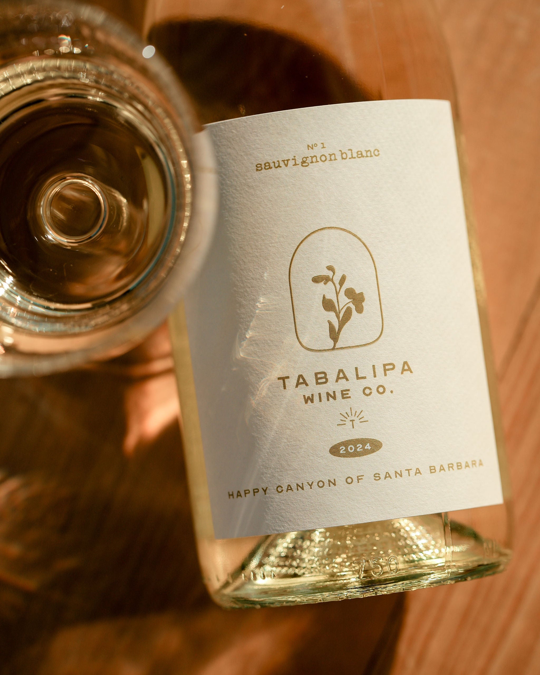Tabalipa Happy Canyon Sauvignon Blanc wine bottle laying down on table with wine glass to the left with the angle facing down parallel to the table