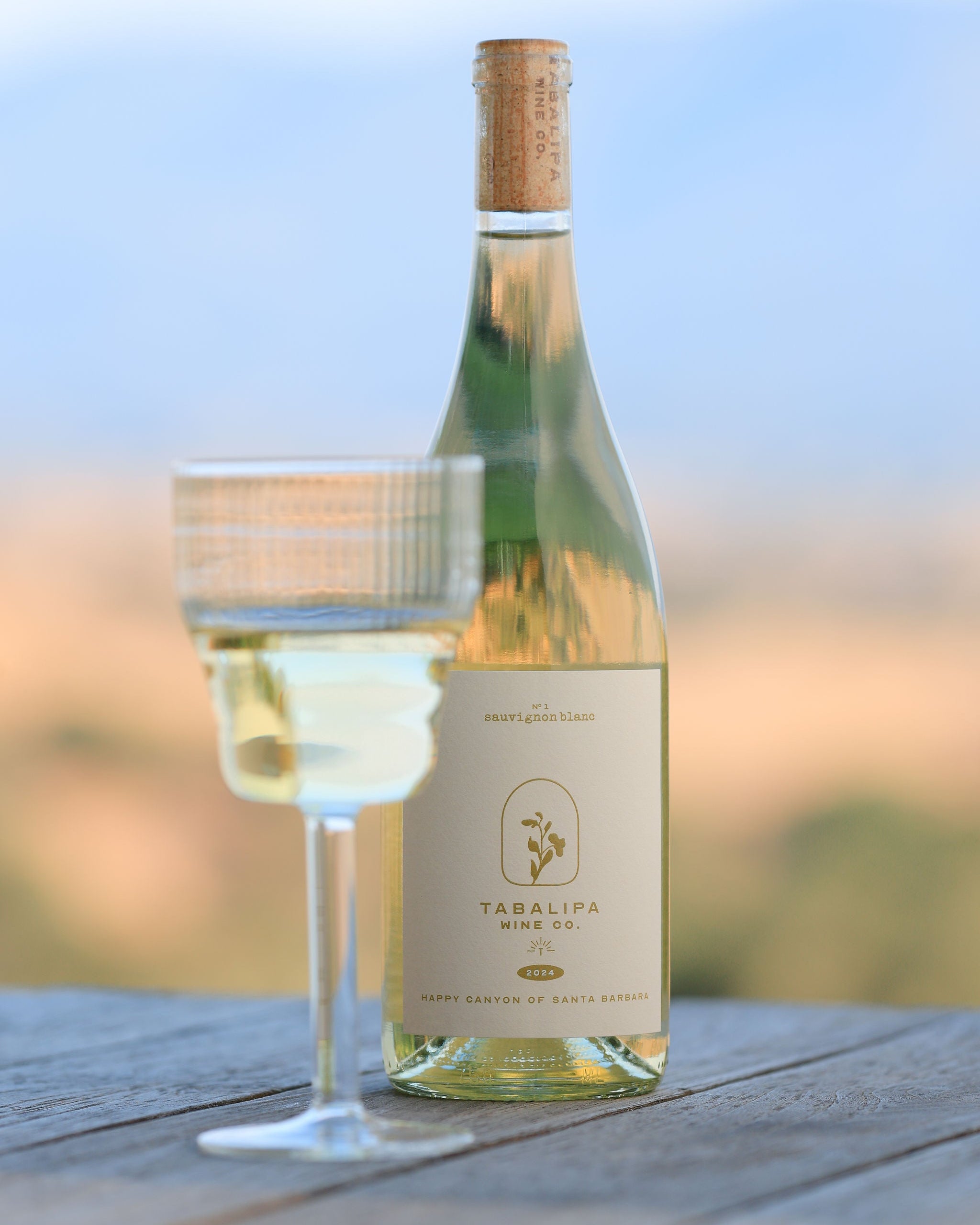Tabalipa Happy Canyon Sauvignon Blanc wine bottle, front label view on rustic table with wine glass to the left just lightly out of focus