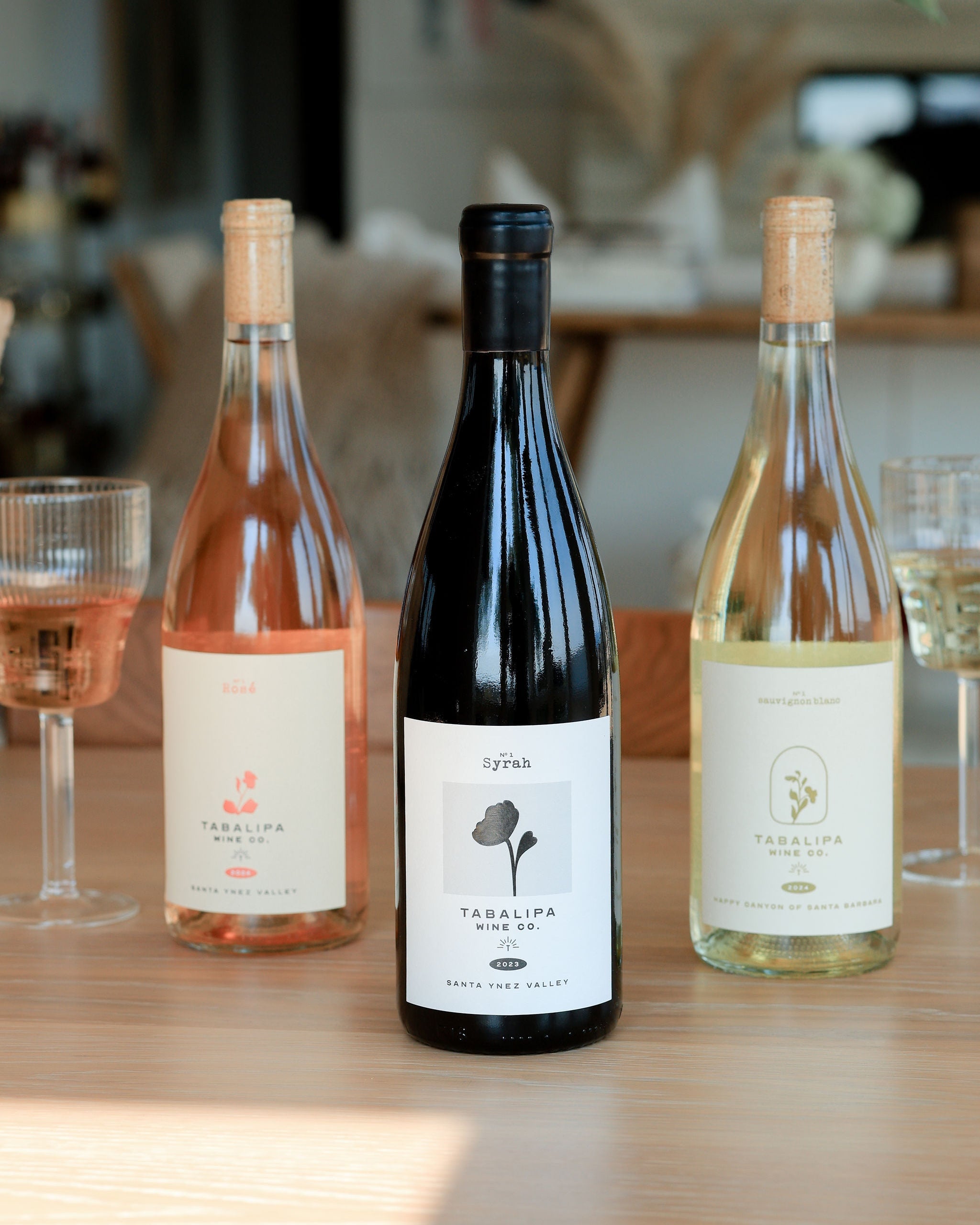 Three bottles of Tabalipa rosé wine lined up on a wooden table, with wine glasses partially visible on both sides in a softly blurred indoor setting.