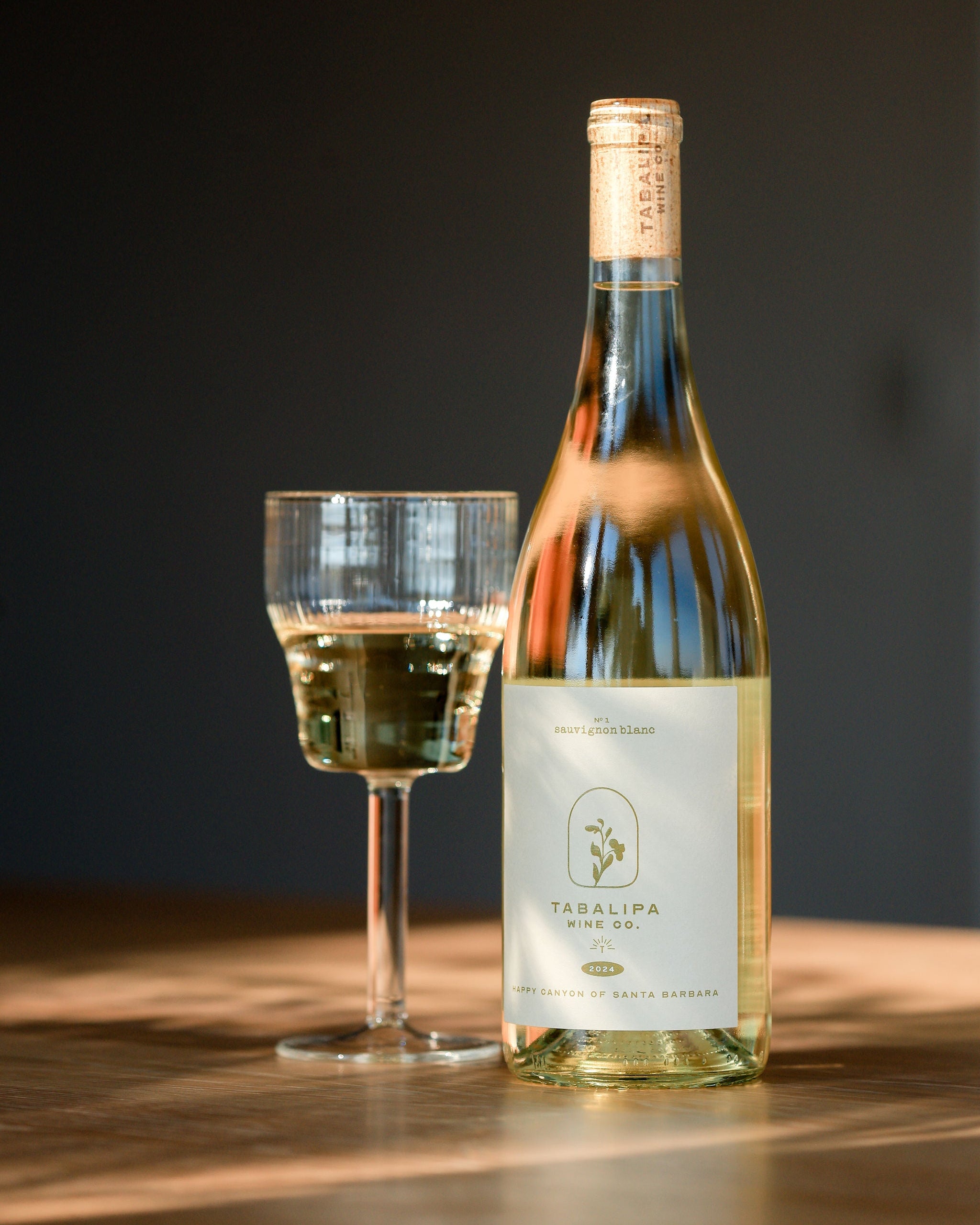 A bottle of Tabalipa White 100% grapevine wine and a glass of the pale off-white wine, set on a wooden table against a dark background.