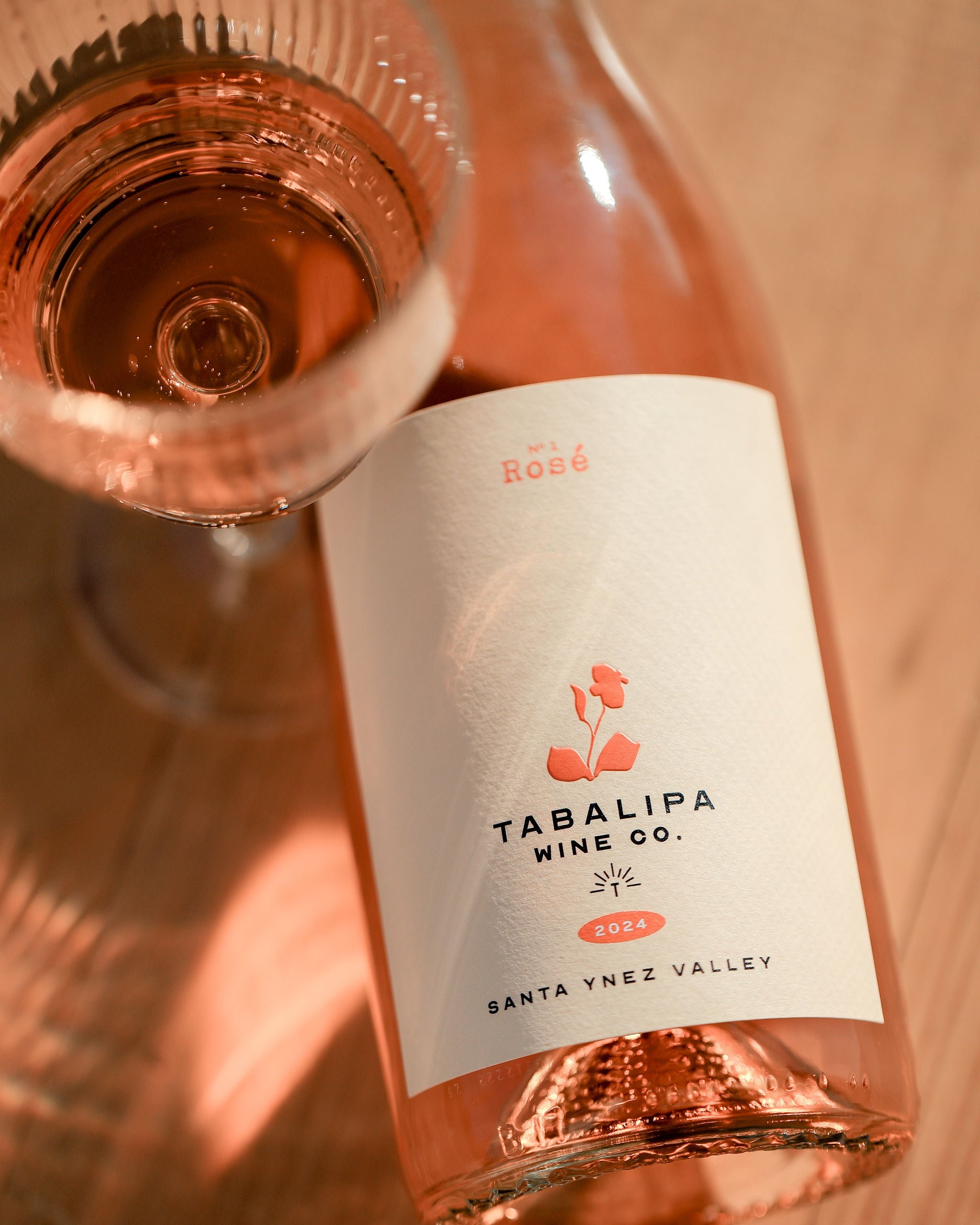 A bottle of Tabalipa Wine Co. rosé wine with a glass of the pinkish rosé liquid, photographed on a warm wooden surface.
