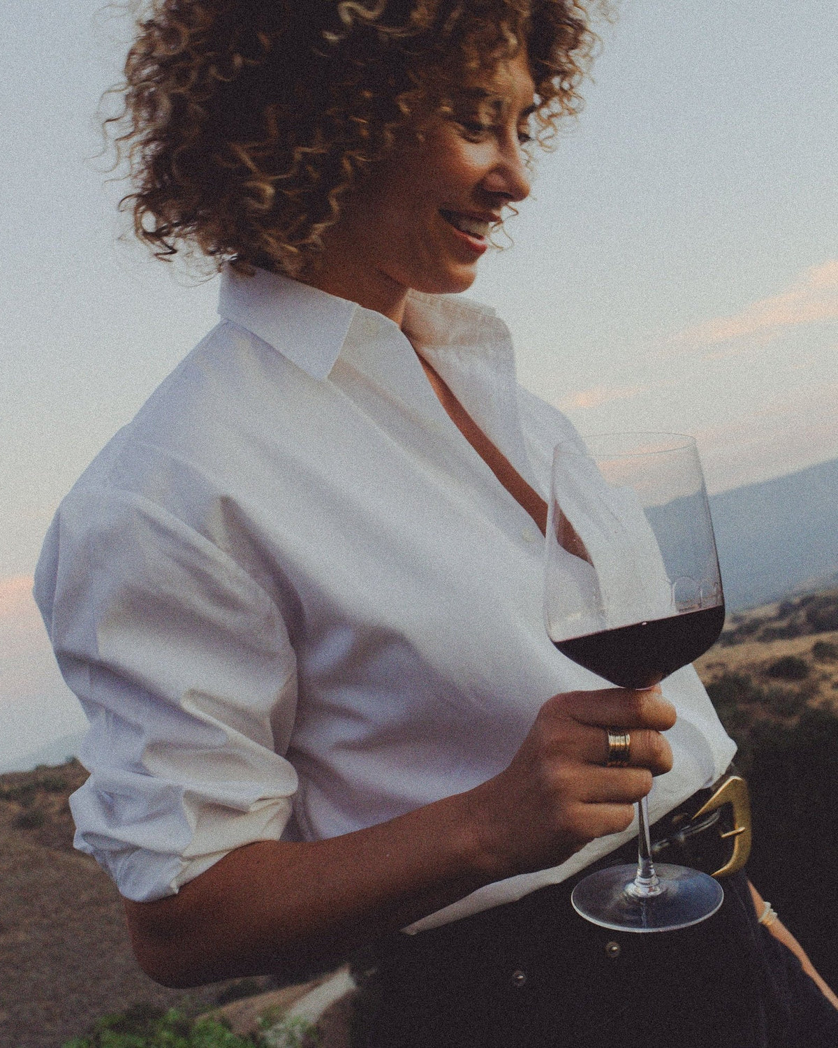 A woman with curly hair holding a glass of red wine, standing in front of a mountainous landscape in Santa Ynez, California.