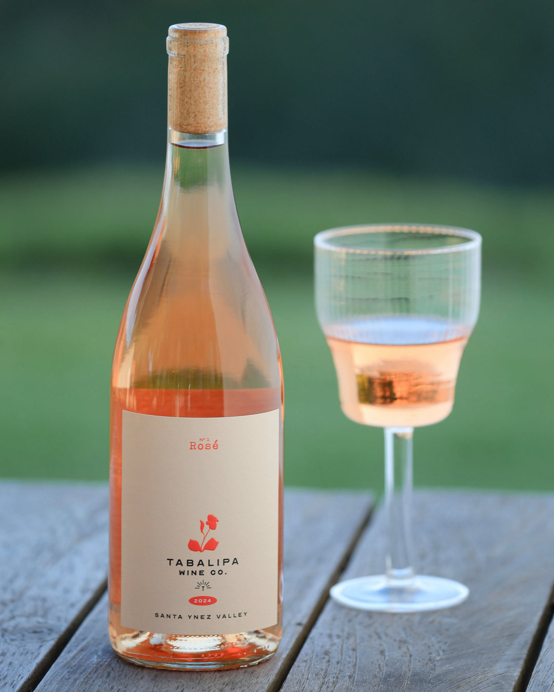 Bottle of rosé with a glass of rose wine to the right of it on a rustic table with an out of focus green background of trees and a grassy field.