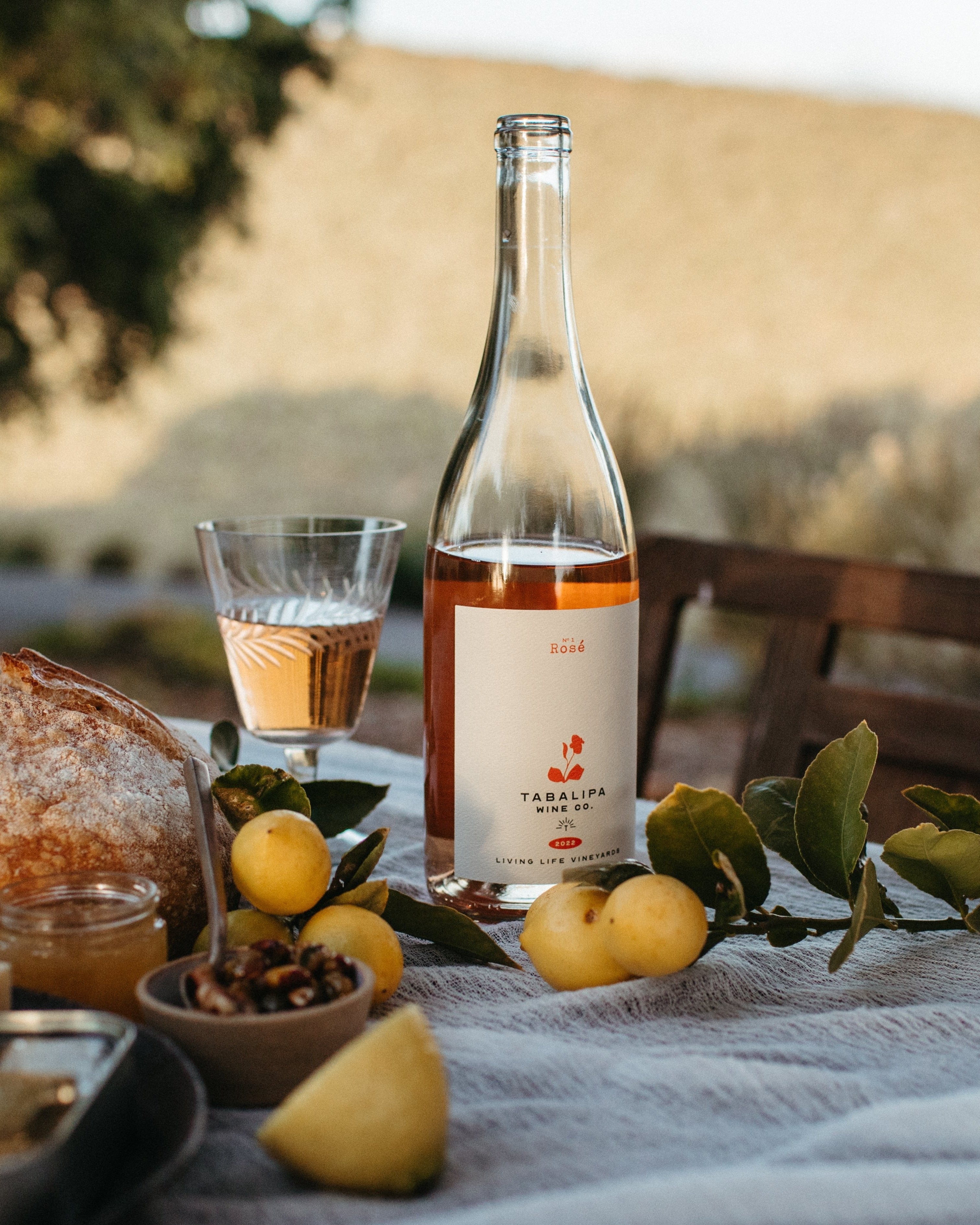 A bottle of rose wine labeled 'No. 1 Rosé' from Living Life Vineyards, displayed on a table with a glass of wine, bread, and fruits.