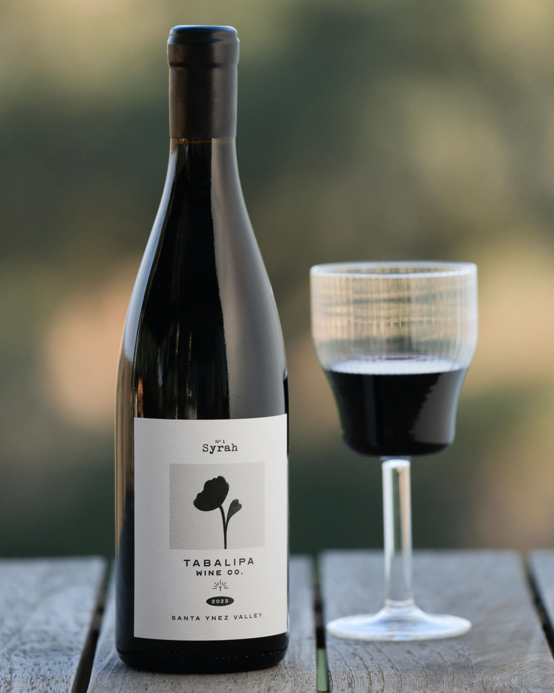 Tabalipa Estate Syrah 2023 wine bottle on rustic table with glass of it to the right of the bottle.