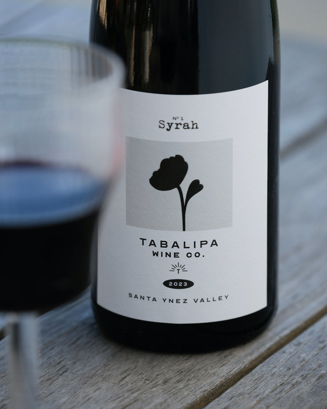 Tabalipa estate syrah on rustic table with a out of focus glass of syrah wine to the right of it.