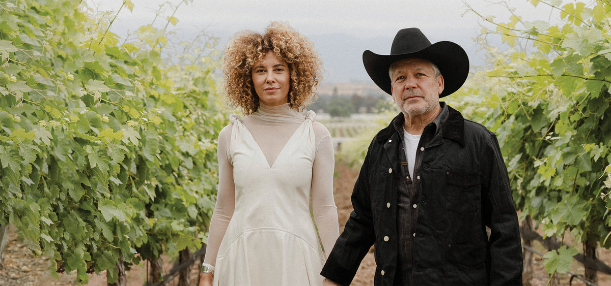 Woman with curly hair and a white dress holding hands with a scruffy man wearing a black cowboy hat and black jacket over a denim shirt with a white t-shirt peeking through standing in the middle of their vineyard wit ha hazy background.