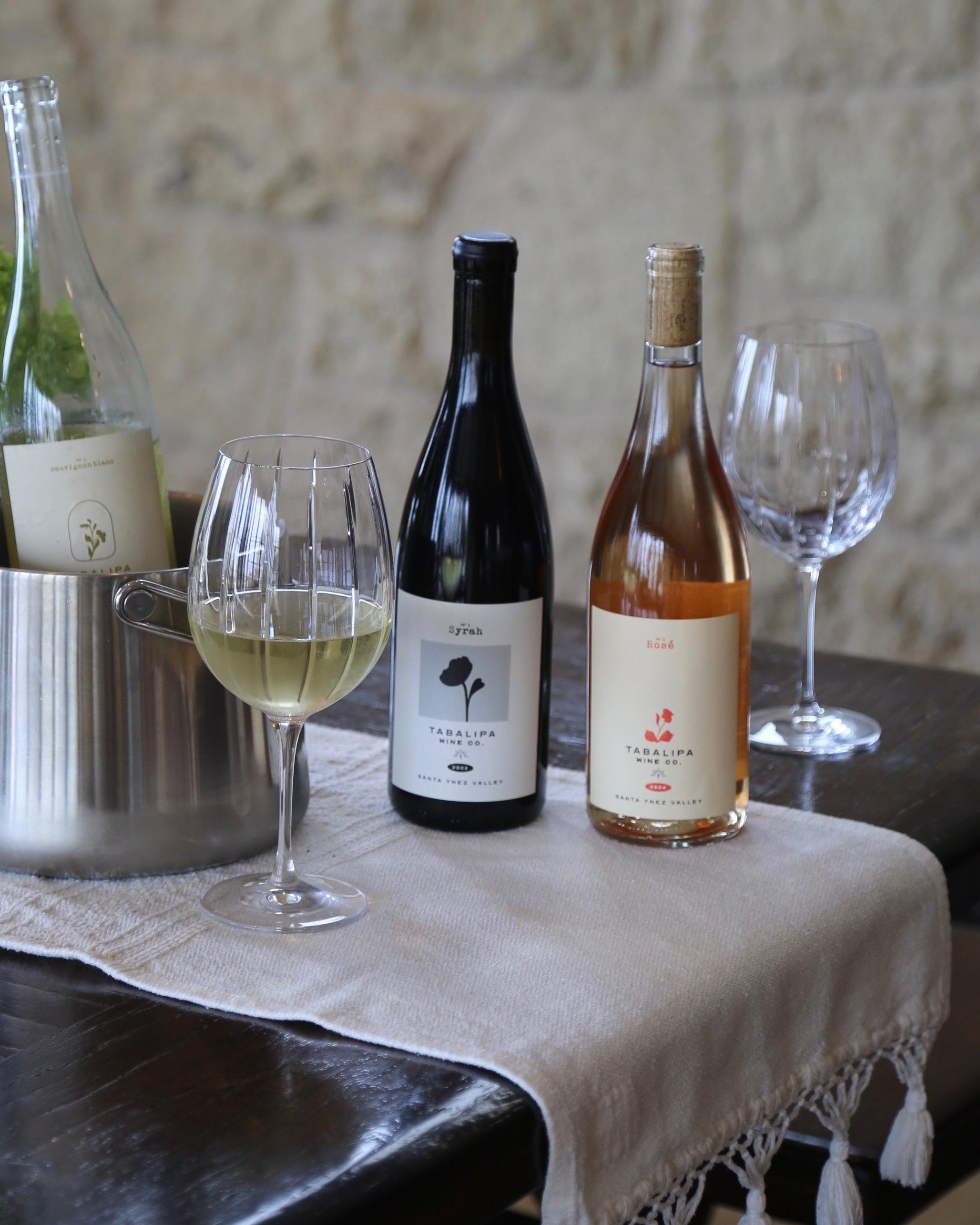 Two wine bottles and two wine glasses arranged on a dark wooden table with a cream woven runner. One bottle is dark with a black label, one is pale pink with a cream label, and a third bottle is partially visible in a metal bucket.