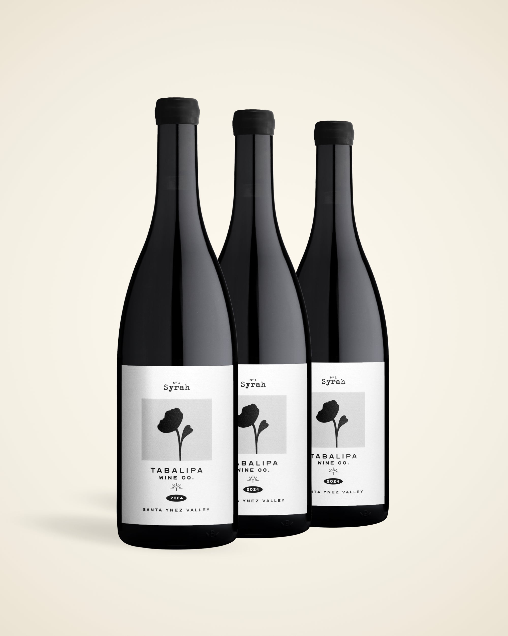 Three dark glass wine bottles with black caps and white labels featuring a black floral graphic, arranged in a row against a light beige background. The labels read 'syria' and 'TABALIPA RED VAR.