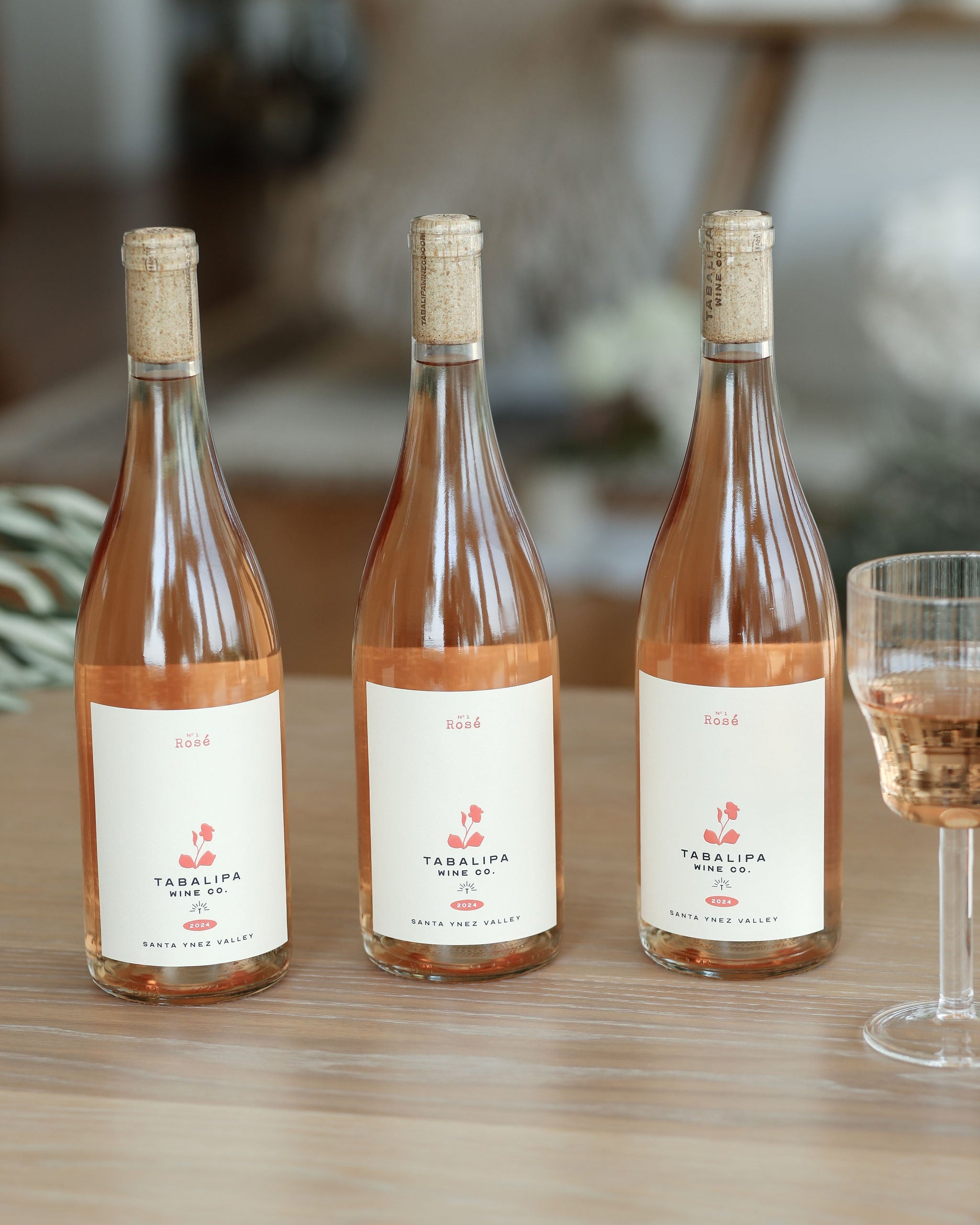 Three bottles of Tabalipa Rosé Red wine with white labels and corks, arranged on a wooden table next to a glass of rosé wine, with a softly blurred indoor background.