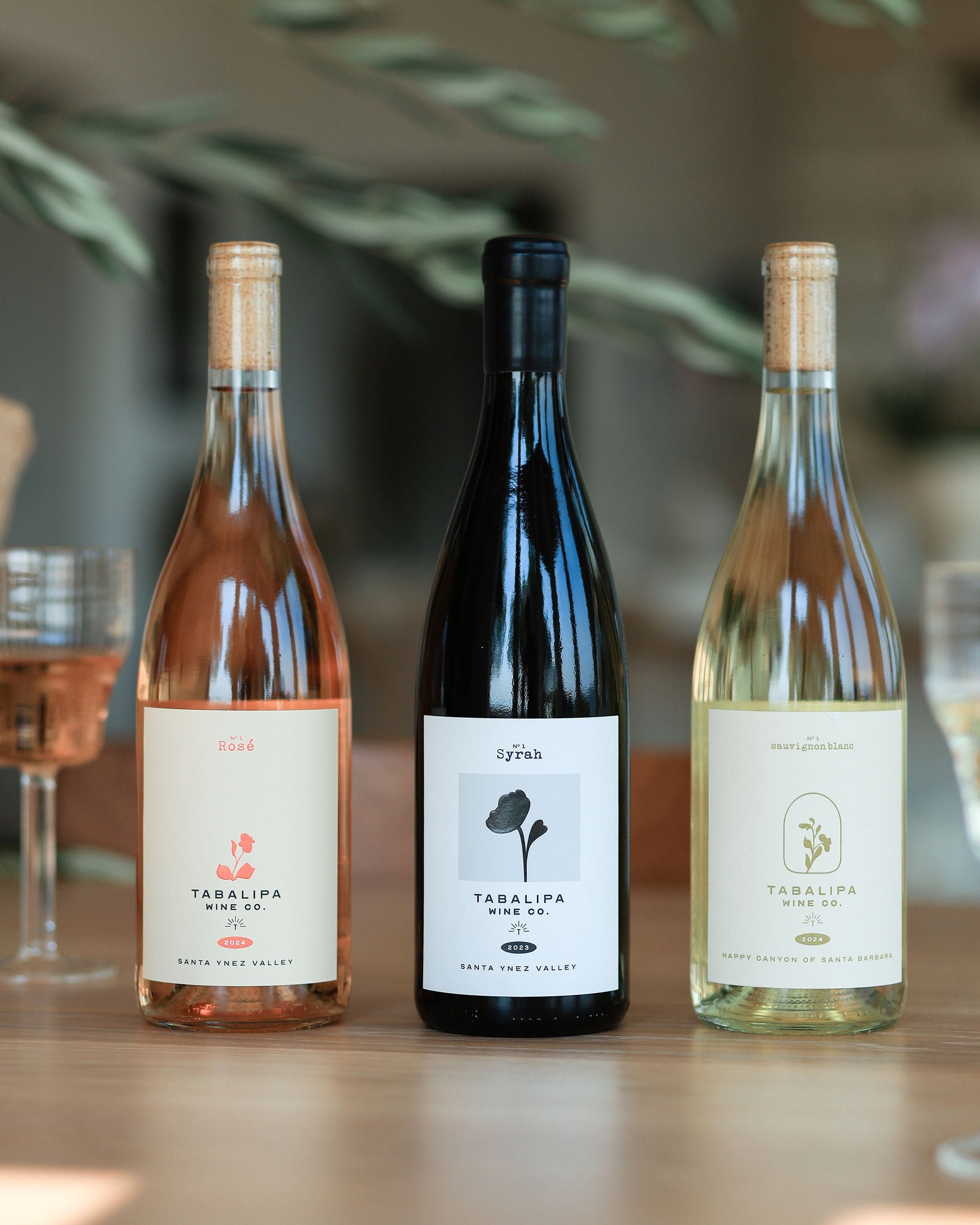 Three wine bottles from Terlina Wine Co. on a wooden table with glasses and greenery in the background.
