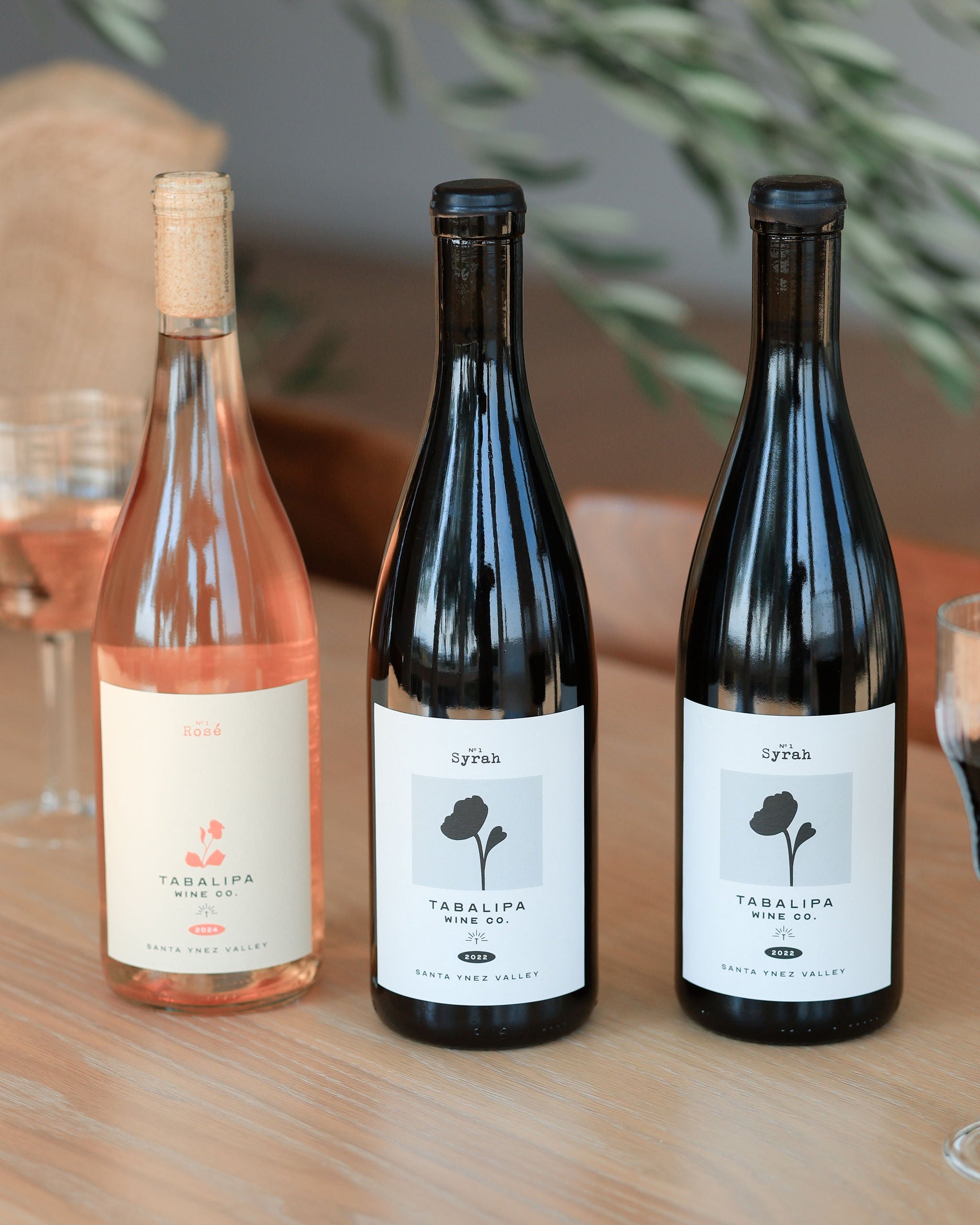 Three bottles of Taralipo wines on a wooden table with glasses and greenery in the background.