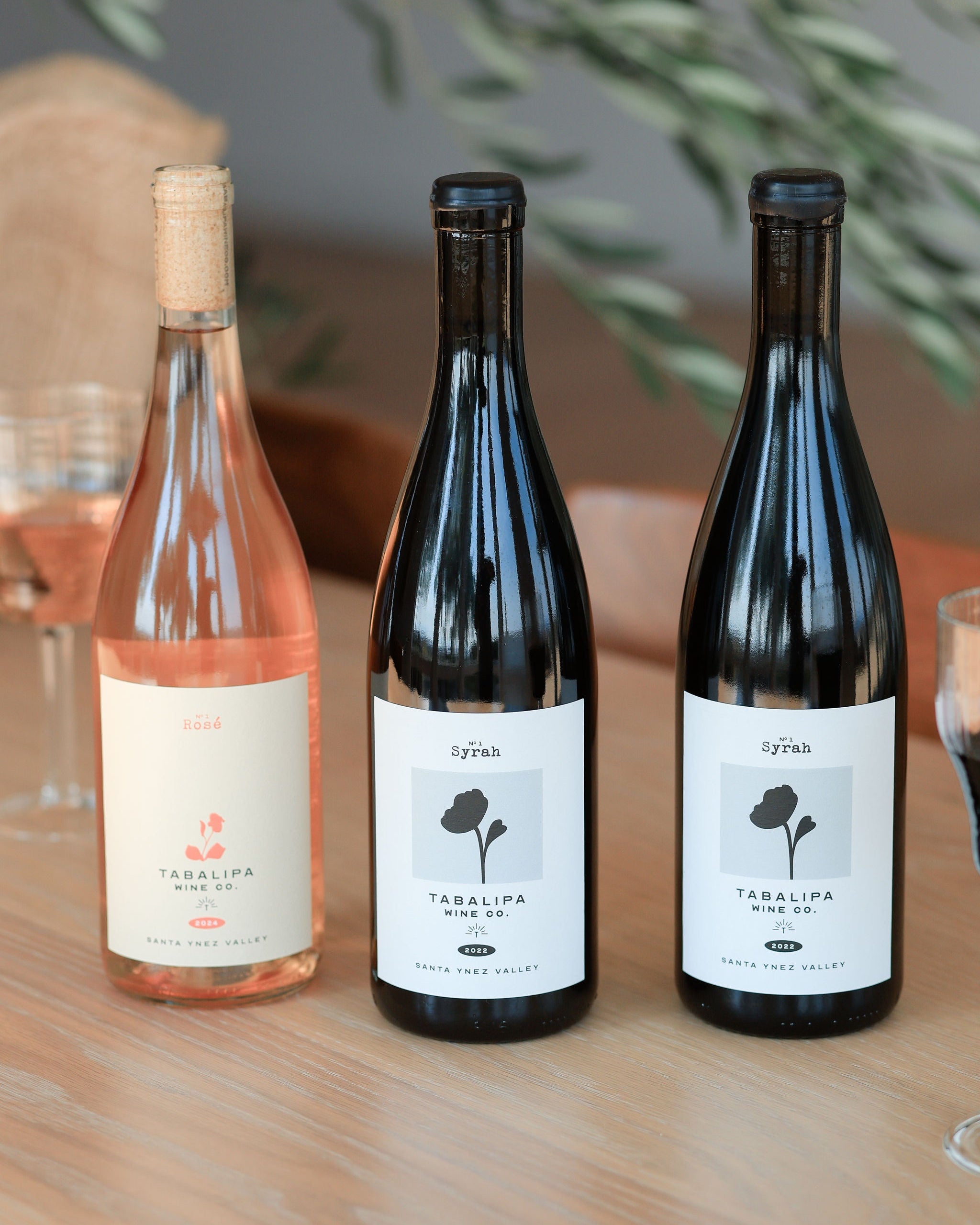 Three wine bottles from Syrah on a wooden table, including a rosé wine bottle and two dark red wine bottles, with wine glasses partially visible in the background.