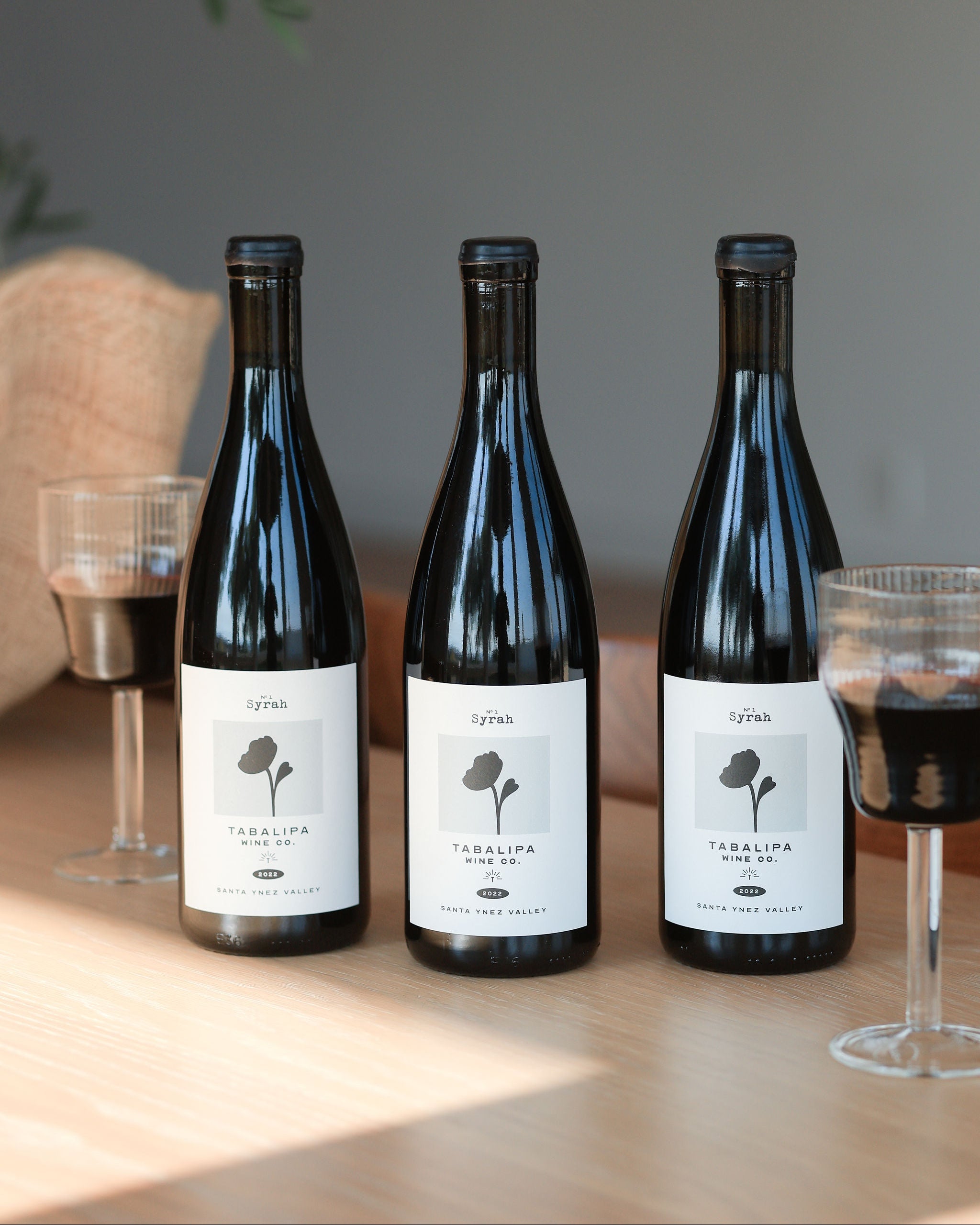Three wine bottles with labels on a wooden table with glasses of red wine.