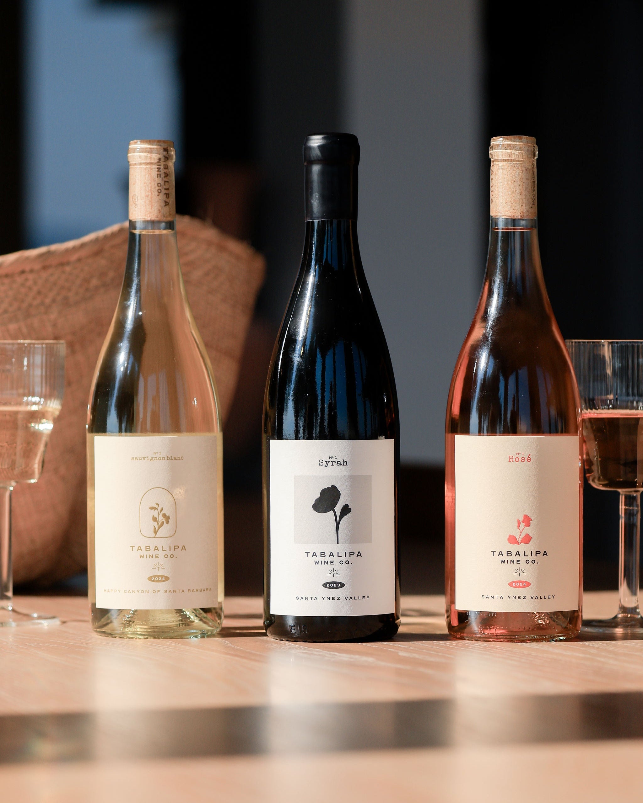 Three wine bottles from Taralupa Winery lined up on a wooden table with wine glasses, featuring a white wine bottle, a dark red wine bottle, and a rosé wine bottle.