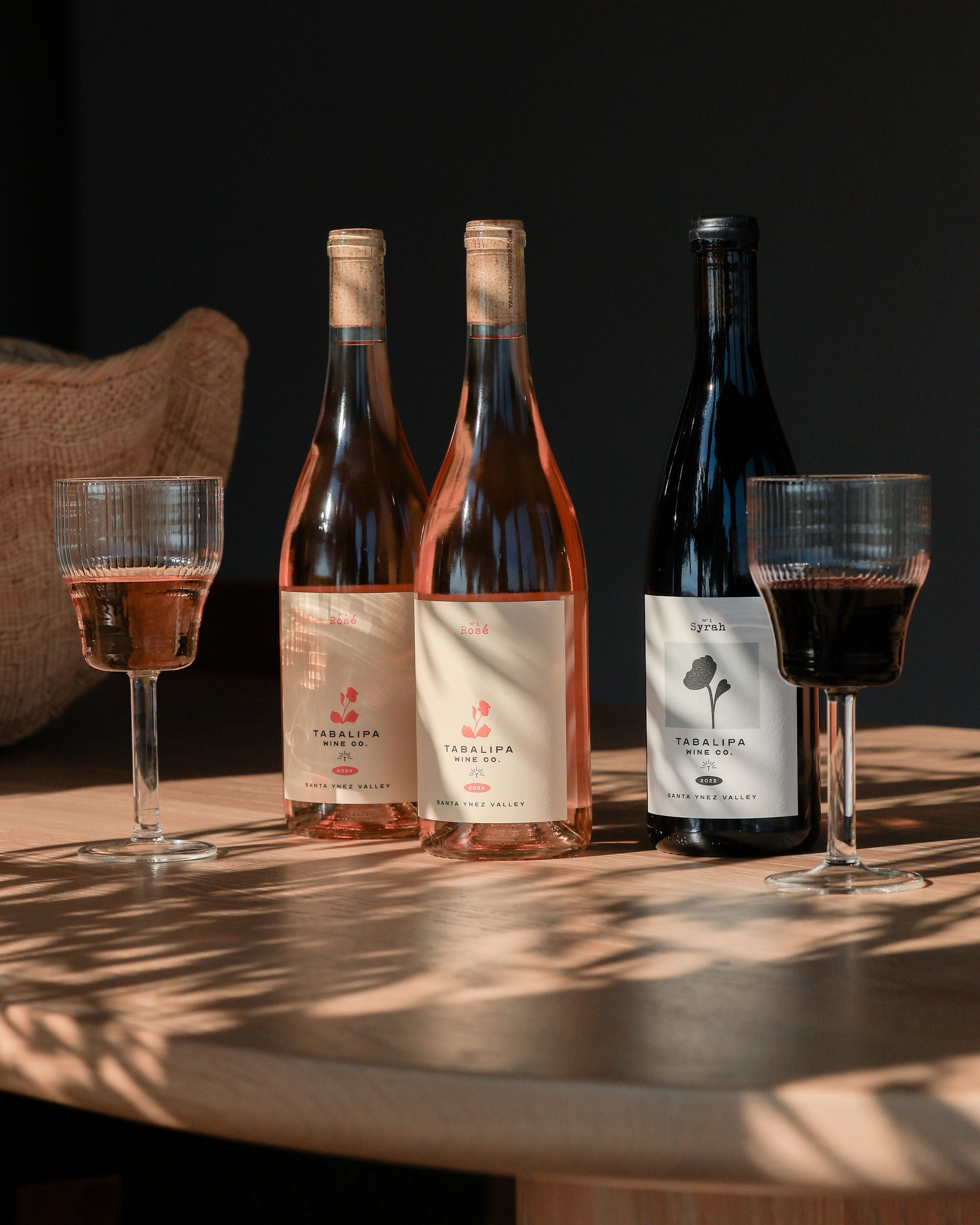Three wine bottles with their labels facing the camera, accompanied by two glasses of wine on a wooden table.