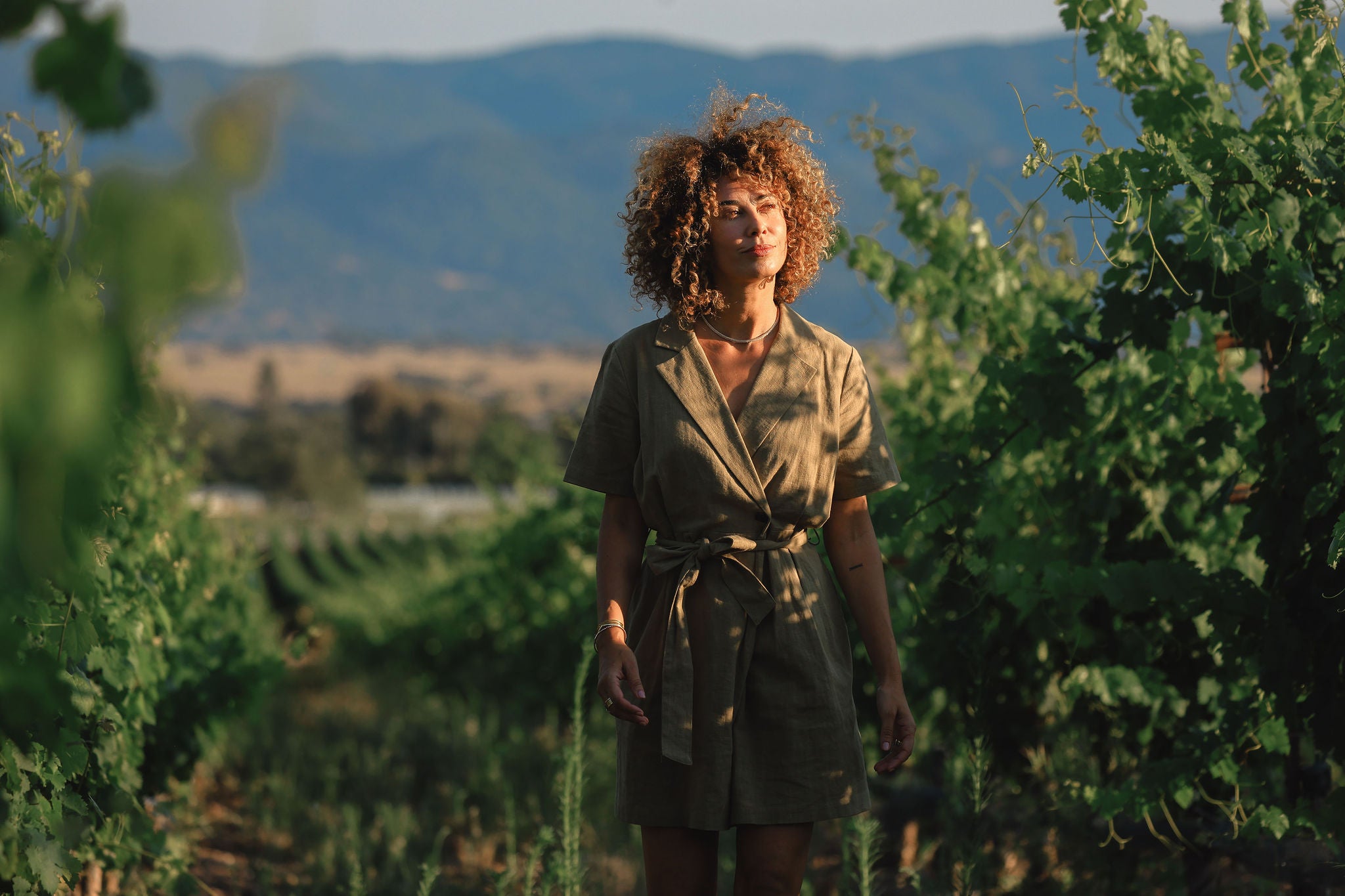 Woman standing in a vineyard wearing a olive green dress with curly hair with mountains in the background