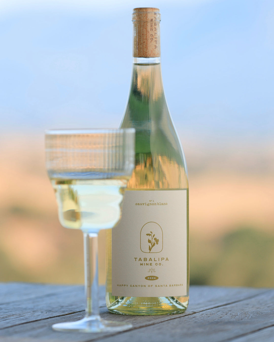 Bottle of Tabalipa Sauvignon Blanch with a glass of white wine on a rustic wooden table with a blurred background of yellow grass and green trees.