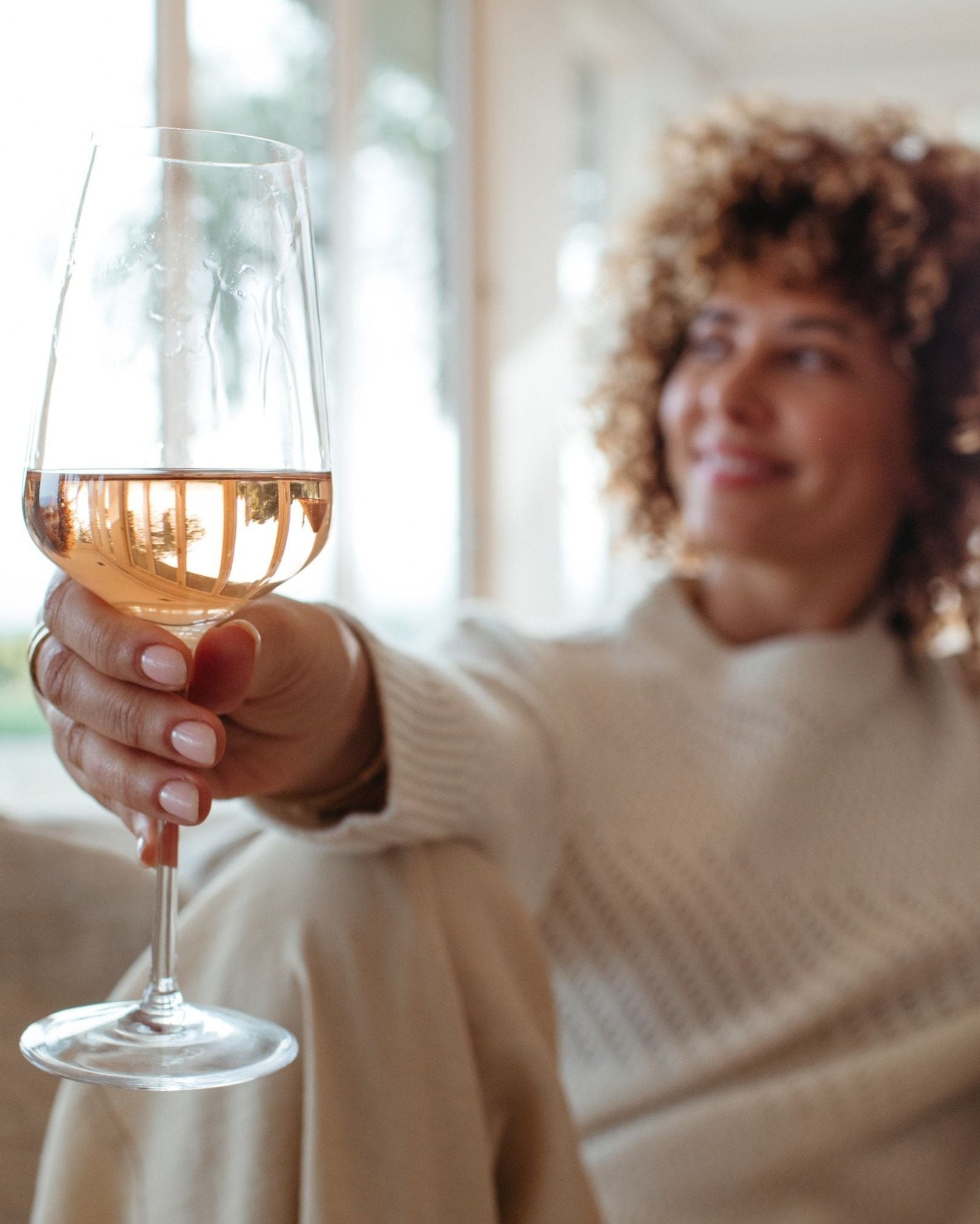 A woman wearing a chunky sweatshirt with curly hair holding a glass of rose wine with a smile on their face, sitting in a well-lit room.