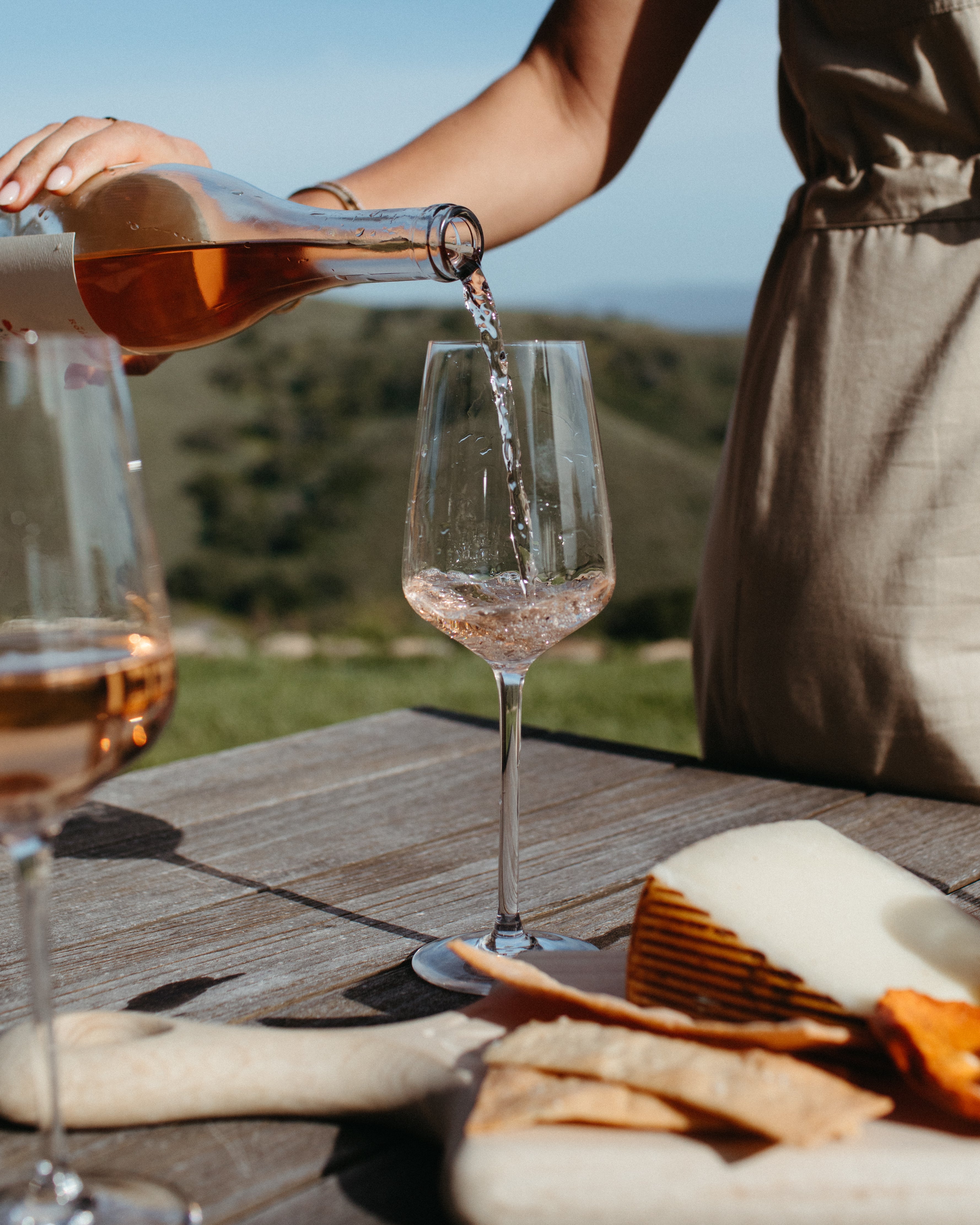 A woman in a olive green jumpsuit pouring rose wine into a glass on a rustic table outside with a cheeseboard with crackers with a scenic background.
